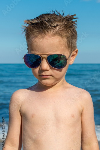 a beautiful little boy posing on a beach by the sea with sunglasses