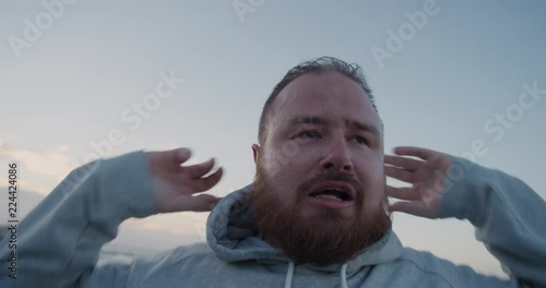 young overweight runner man resting takes off hood exhausted after intense running exercise training difficult weight loss challenge on seaside at sunrise close up