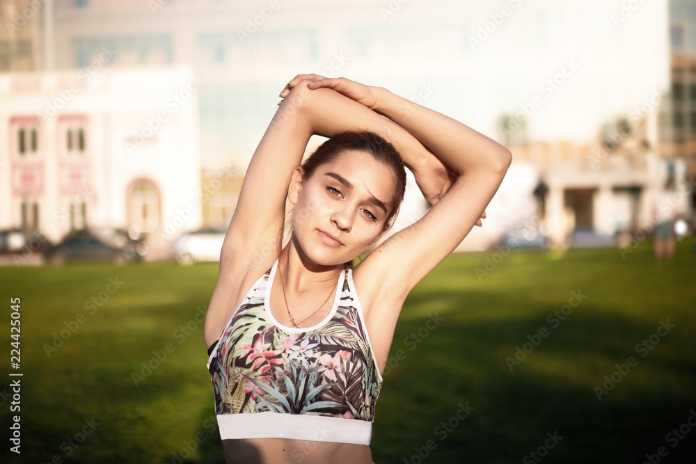 Sports fitness slim woman in leggings with tropical print make stretching exercises looking camera outdoors in the park on green grass with sundown light.