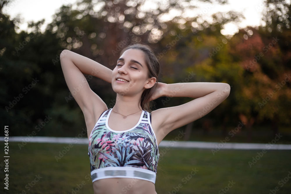 Beautiful brunette sports fitness slim woman in top with tropical print make stretching exercises with eyes closed outdoors in the park on green grass with sundown light. Beautiful brunette sports fitness slim woman in top with tropical print make stretching exercises with eyes closed outdoors in the park on green grass with sundown light.
