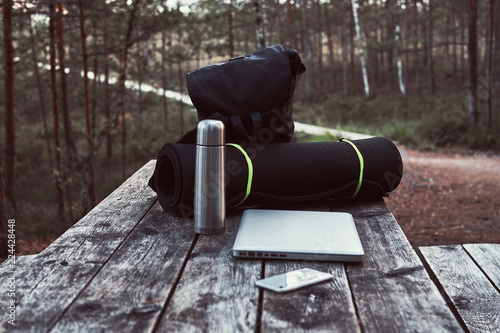 Travel concept. Backpack, tourist mat, laptop, smartphone and thermos bottle on a wooden table in beautiful forest.