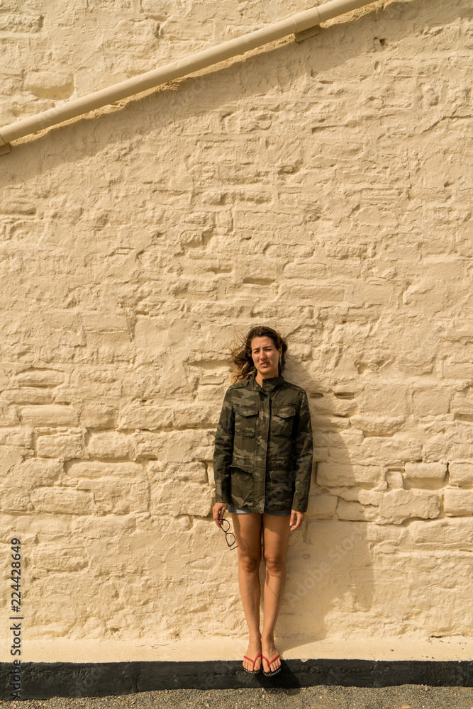woman in military jacket holding her glasses and posing in front of a stylish wall