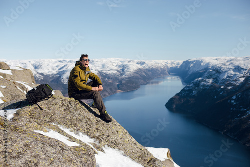 Top view of happy traveler man sitting on a rocky surface on a cliff and enjoying the scenic view of the fjord and mountains. Preikestolen, Pulpit rock, Lysefjord, Norway