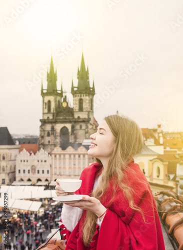 Charming smiling young woman in red blanket with tea sitting in Europe city centre. Expressing positivity, waiting for christmas holidays, smiling to side. Cozy photo of traveling girl in Prague.