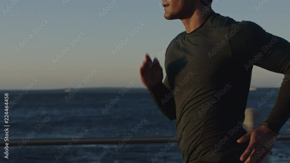attractive young man running athlete endurance runner training jogging cardio exercise workout on calm ocean seaside run wearing earphones at sunset slow motion