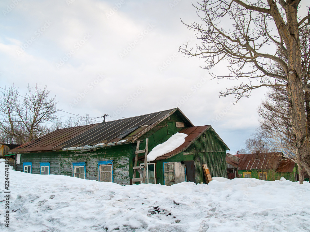 A green wooden house with blue platbands on the windows stands covered with snow in winter