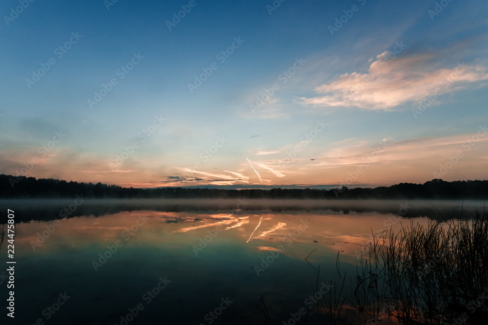 Beautiful, red dawn on the lake. The rays of the sun through the fog. The blue sky over the lake, the morning comes, the sky is reflected in the water.