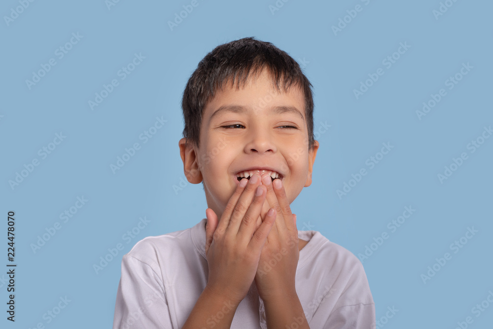 Close up emotional portrait of little boy wearing a white shirt on blue background in studio. He is laughs with his hands over his mouth Close up emotional portrait of little boy wearing a white shirt on blue background in studio. He is laughs with his hands over his mouth