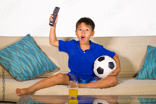 lifestyle portrait at home of young 7 or 8 years old boy holding soccer ball watching excited and nervous football game on television sitting at living sofa couch