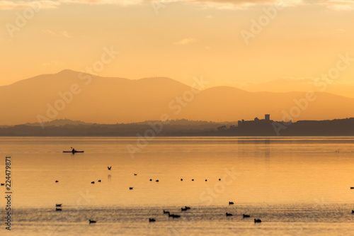 Beautiful view of Trasimeno lake  Umbria  Italy  at sunset  with orange tones  birds on water  a man on a canoe and Castiglione del Lago town on the background