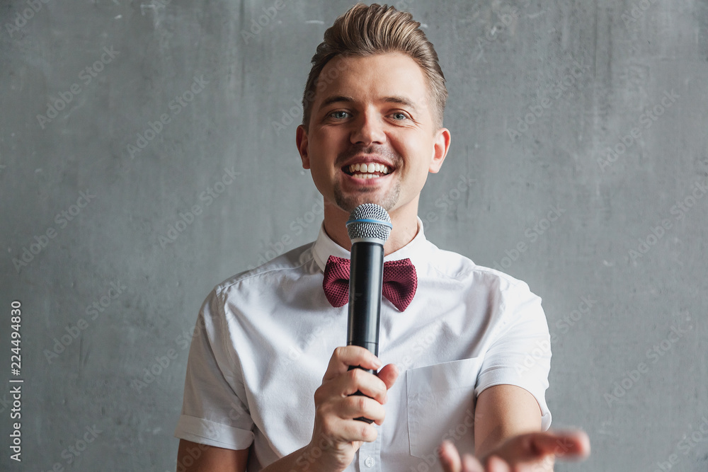 A young attractive funny joyful emotional man winks in a white shirt and a butterfly with a microphone in his hands, on a gray background A young attractive funny joyful emotional man winks in a white shirt and a butterfly with a microphone in his hands, on a gray background