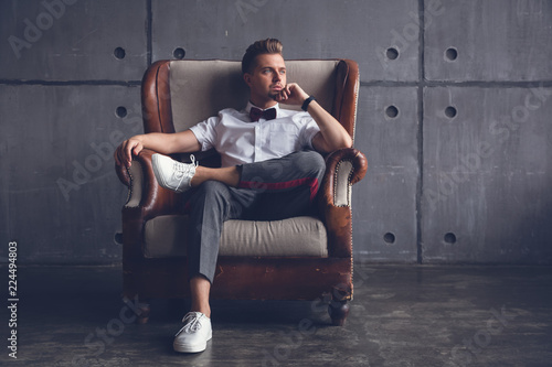 Young attractive serious glamorous male hipster in white shirt and butterfly sitting in chair, on gray background