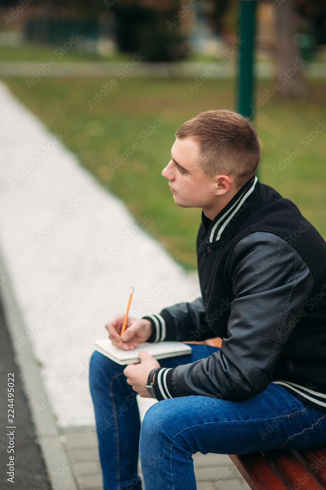 Student in a black jacket sits in a park on a bench writes down his thoughts in a notebook. Handsome boy