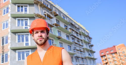Builder in orange vest and helmet works construction site. Safety of builder at work concept. Guy in protective helmet stand in front of building made out of red bricks. Control construction process