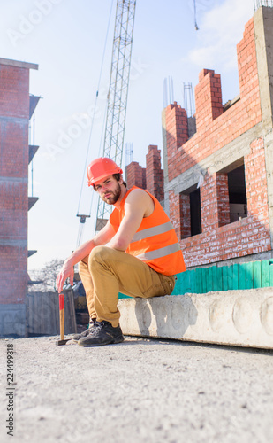 Builder vest and helmet construction site sit relaxing. Guy protective helmet sit in front of building made out of bricks. Take minute to relax. Man hammer take break working day at construction site