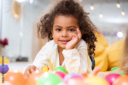 smiling african american kid lying on carpet with toys and looking at camera in kindergarten