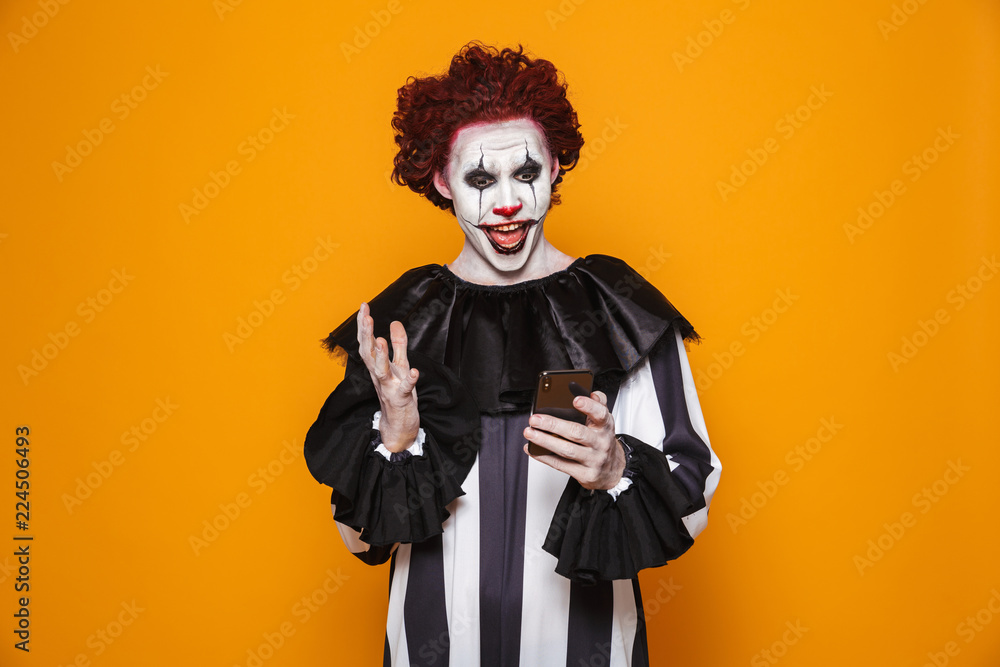 Young clown man 20s wearing black costume and halloween makeup holding smartphone, isolated over yellow background