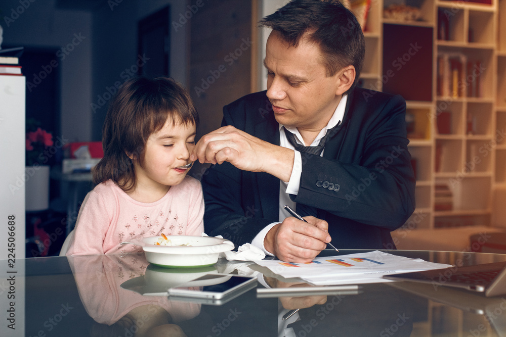 Father in suit working with papers and feeding daughter. Business at home concept. Father in suit working with papers and feeding daughter. Business at home concept.