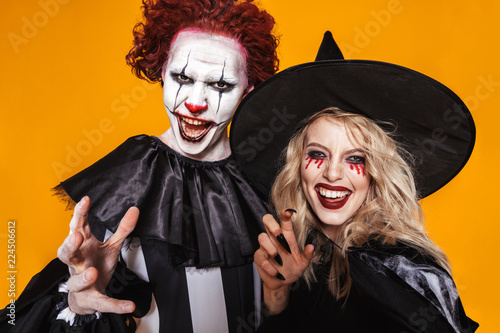 Image of witch woman and clown man wearing black costume and halloween makeup scaring at camera, isolated over yellow background