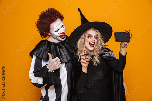 Photo of horrific witch woman and joker man wearing black costume and halloween makeup taking selfie on smartphone, isolated over yellow background