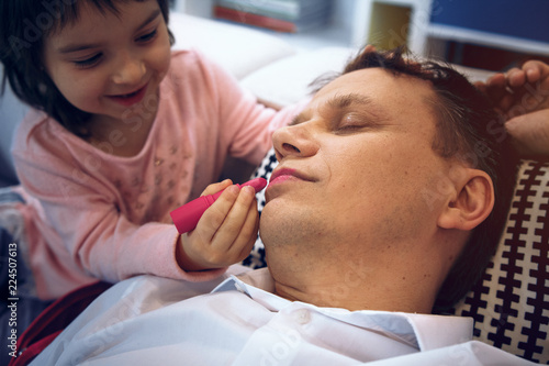 Cute little girl apply lipstick to father while he is sleeping. Spending time happily.