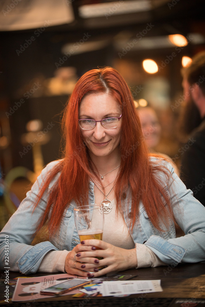 Red-haired young woman in glasses sits in a bar with a mug of beer Red-haired young woman in glasses sits in a bar with a mug of beer