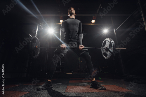 Training with barbell, dark background. Sporty man performs exercise of deadlift.