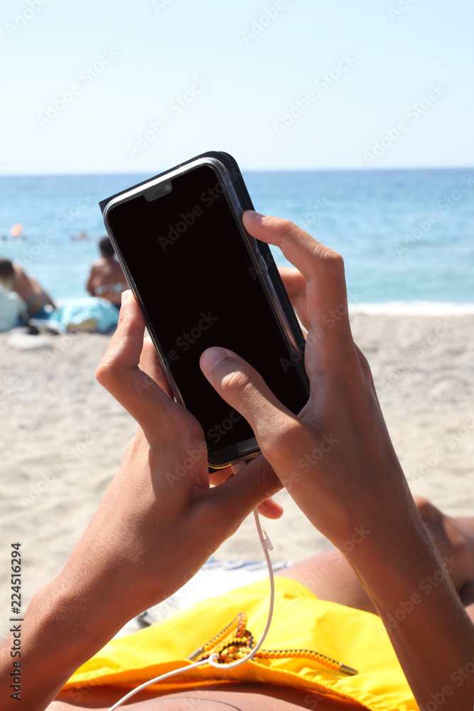 caucasian teenager hand's holding a smartphone lei dow on a sun bed on the beach, clipping path for the screen included