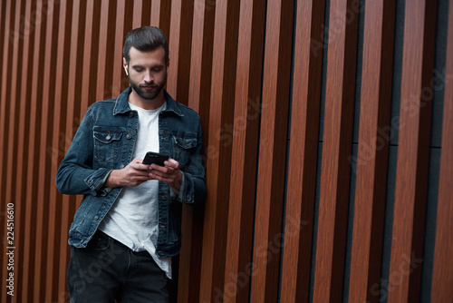 Freestyle. Young man standing leaning on wooden wall isolated browsing internet on smartphone concentrated