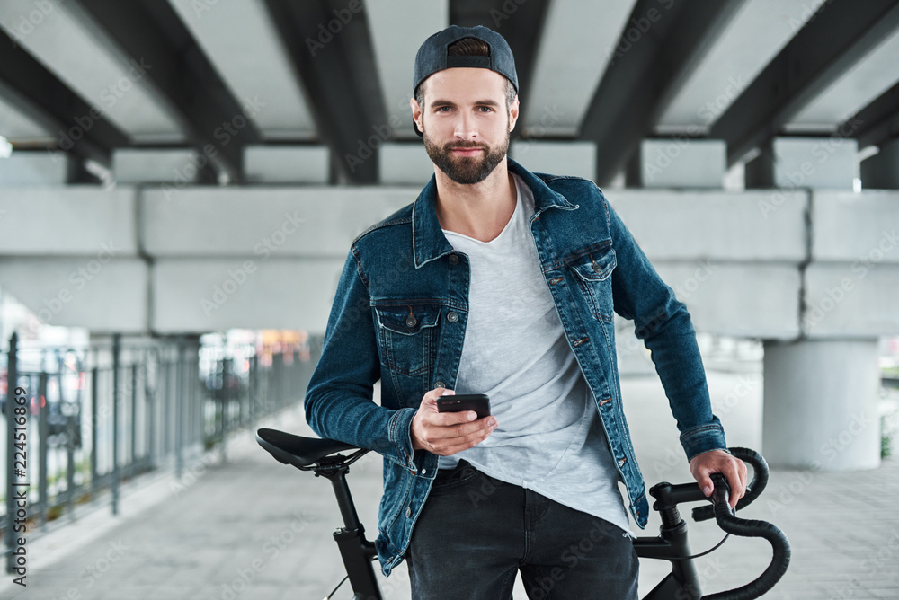 Outdoors leisure. Young stylish man sitting on bycicle on city street holding smartphone looking camera smiling happy