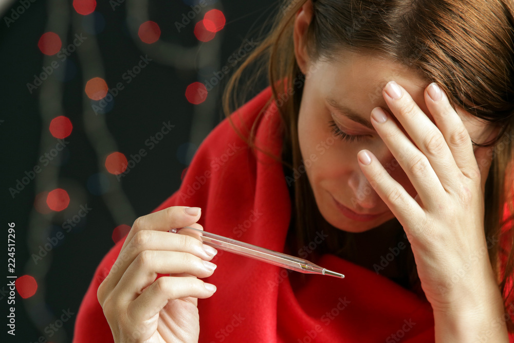 Obraz premium Young woman wrapped in warm red coverlet holds thermometer which shows high temperature, cold and flu during the festive winter holiday season