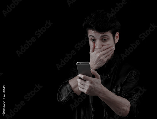 Muscular handsome young man looking at his phone surprisedly with black jacket in front of black background photo