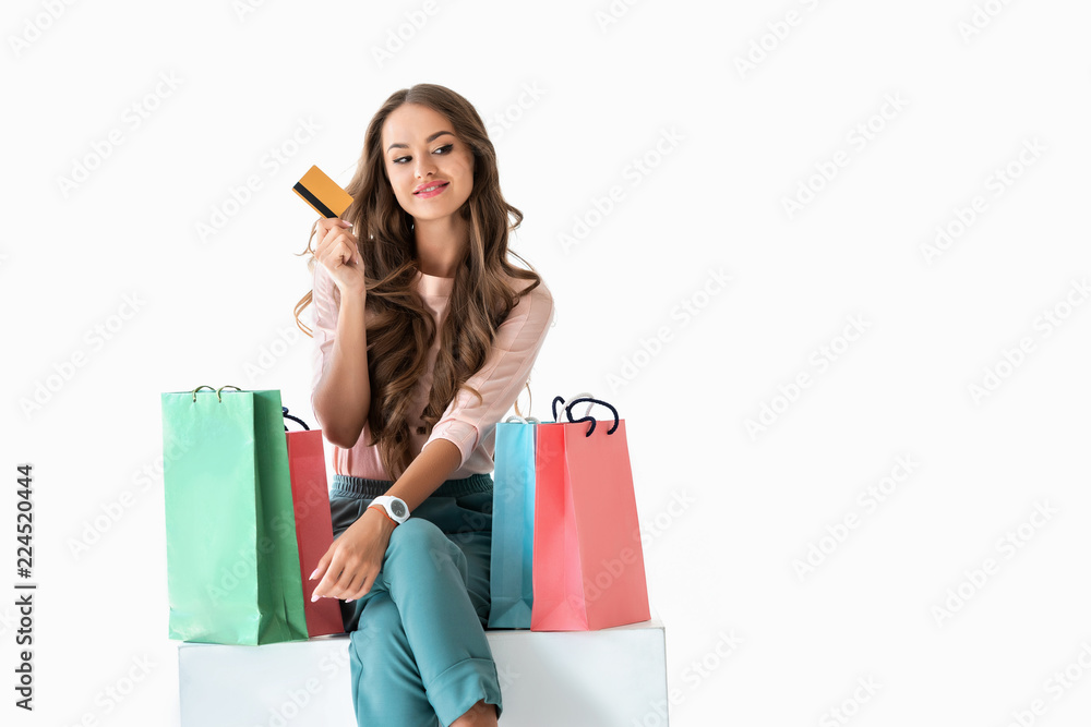 smiling young woman holding golden credit card and sitting on white cube with shopping bags, isolated on white smiling young woman holding golden credit card and sitting on white cube with shopping bags, isolated on white
