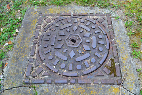 Decorative metal forged sewer hatch cover in green grass, top view close up