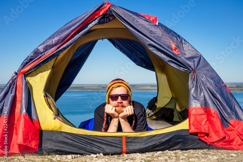 portrait of stylish ginger red-haired beard tourist man in a colorful hat made of yak wool from Nepal lying down in tent wrapped in a sleeping bag the background of the tent and the lake photo