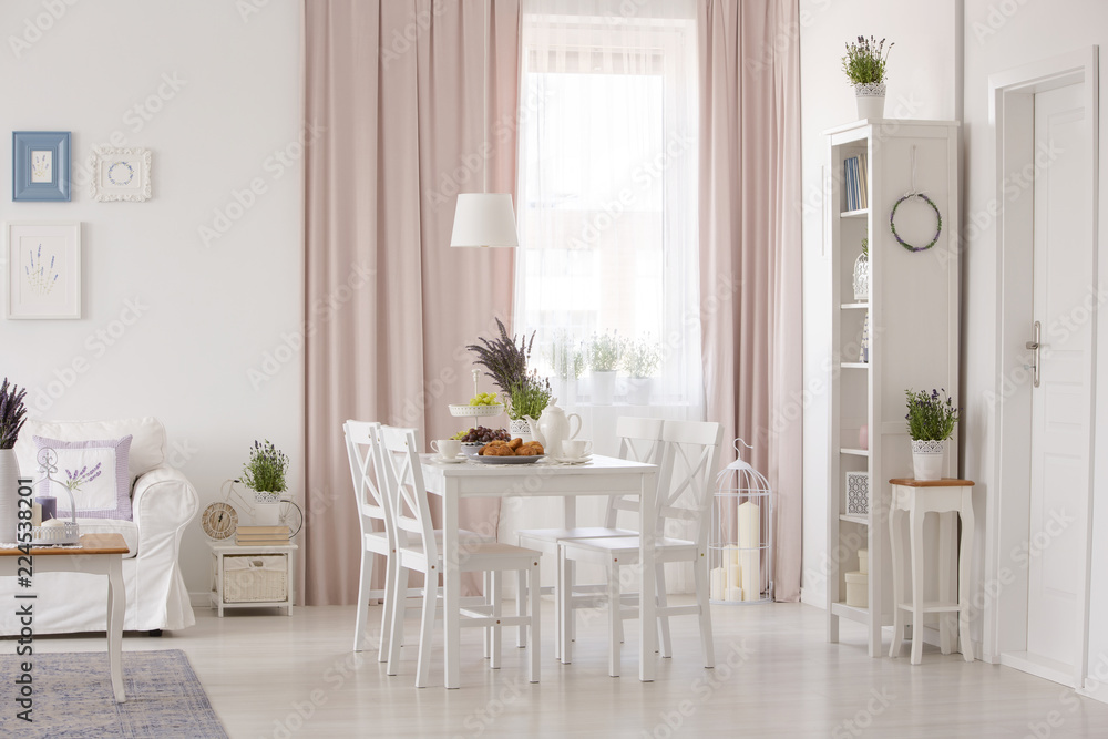 Fototapeta premium White chairs at table near couch in pink apartment interior with drapes and plants. Real photo