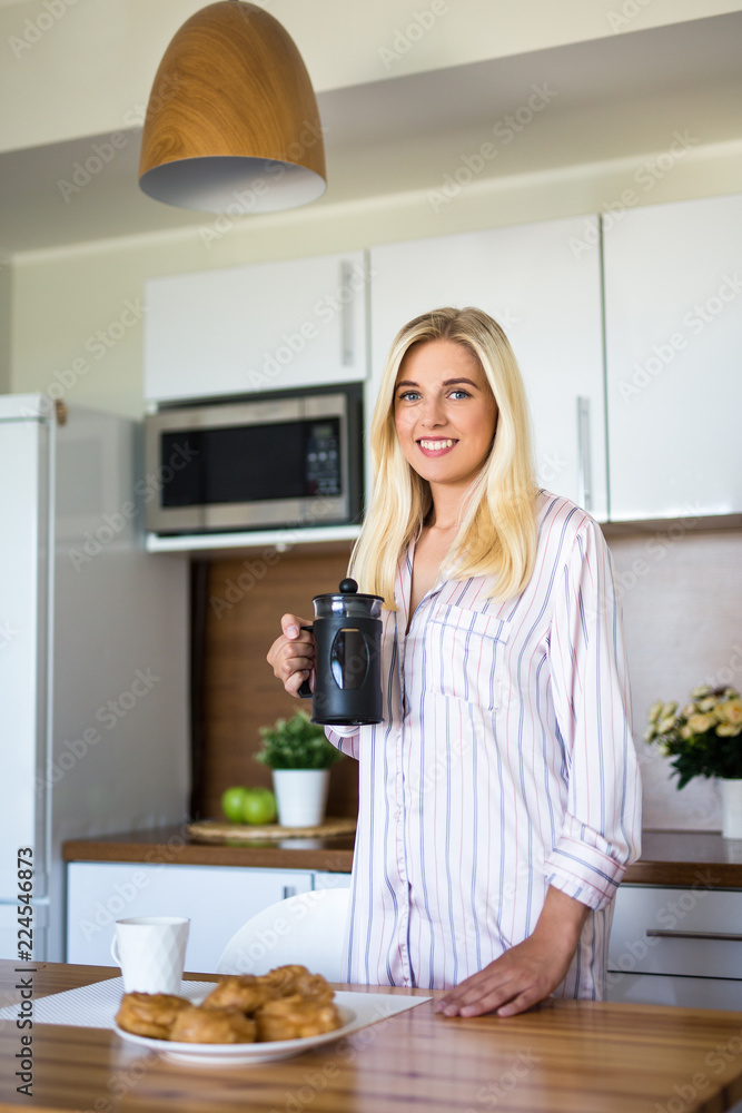breakfast concept - portrait of young woman in pajamas with french coffee press in modern kitchen