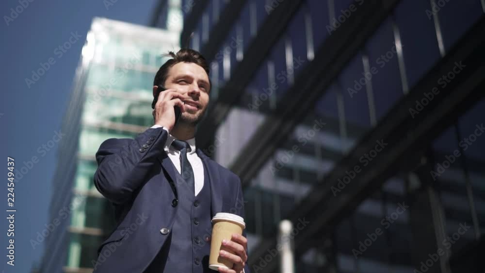 Confident young businessman walking with coffee to go in street with skyscrapers on a summer day and stopping to talk on the smartphone. Tracking slow motion low angle shot Confident young businessman walking with coffee to go in street with skyscrapers on a summer day and stopping to talk on the smartphone. Tracking slow motion low angle shot