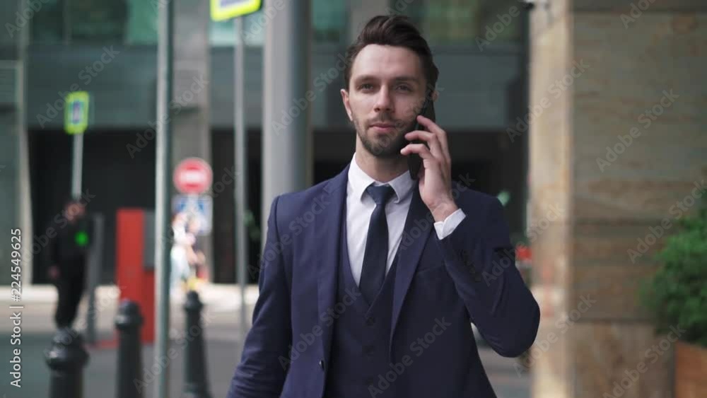 Hansome young caucasian businesman in suit and tie walking in street and talking on smartphone. Tracking slow motion medium shot Hansome young caucasian businesman in suit and tie walking in street and talking on smartphone. Tracking slow motion medium shot