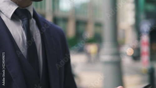 Hands of young caucaisian businesman in a dark suit swiping across his cell phone screen while he is standing outside on a summer day. Tilt up slow motion medium shot photo