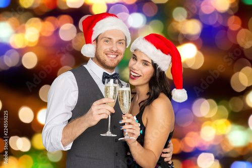 celebration, people and holidays concept - happy couple in santa hats with glasses of non alcoholic champagne at christmas or new year party over festive lights background