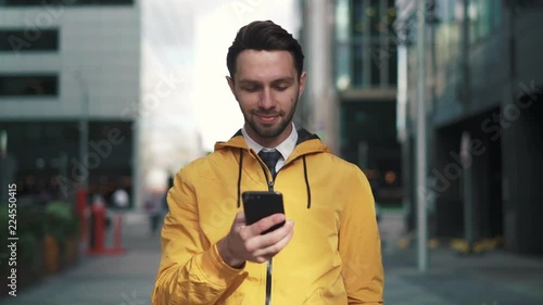 Hansome young caucasian man with fair hair and beard wearing yellow jacket walking in street, dialing a number, smiling and starting to talk on smartphone. Tracking slow motion medium shot