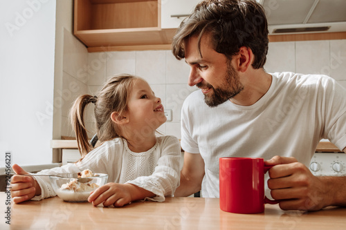 Family. Food. Parenthood. Dad and his little daughter are talking and smiling while having a breakfast in the kitchen at home