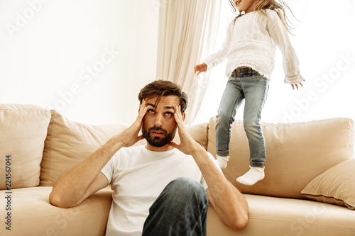 Parenthood. Family. Tired. Dad is having a headache while his little daughter is jumping on the couch at home.