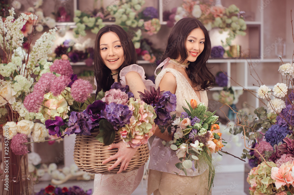Beautiful asian florist girls holding bouquet and basket of flowers for sale against floral bokeh background in flower shop indoors. Two attractive asian females florists working in retail store. 2