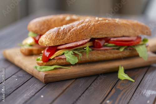  Two baguette sandwiches with salami, cheese, arugula salad, tomatoes and onion on a cutting board. Sandwiches on dark wooden background. Tasty snack.
