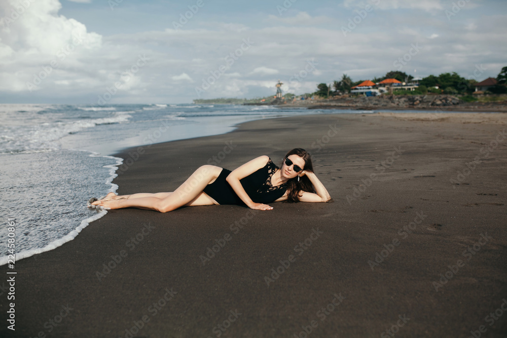 Fashion shot of beautiful girl in black swimsuit and sunglasses lying on the beach with black sand. Bali,Canggu,Indonesia Fashion shot of beautiful girl in black swimsuit and sunglasses lying on the beach with black sand. Bali,Canggu,Indonesia