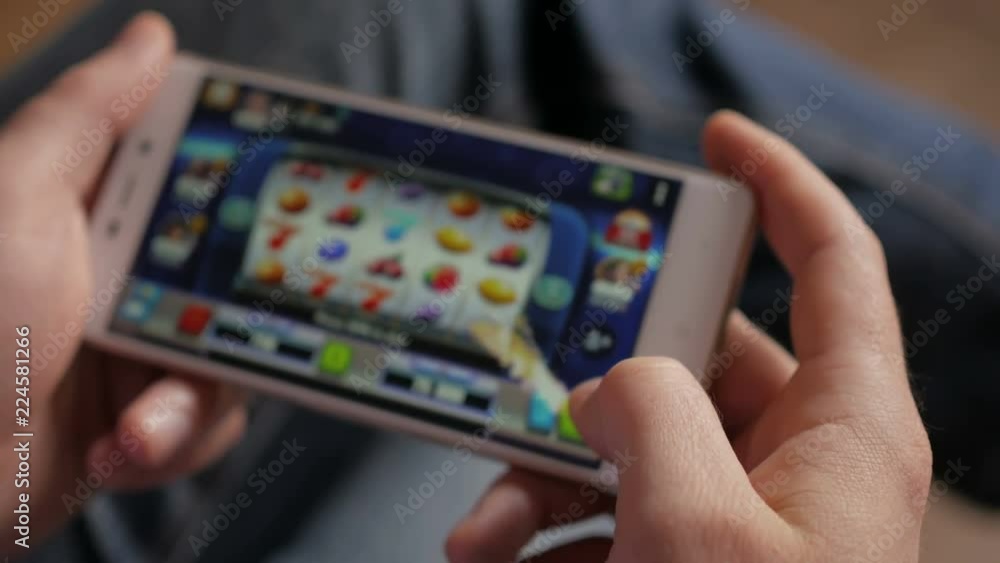Casino slot machine app on mobile screen against hand of man using smartphone. Gambling addicted man in front of online casino slot-machine on mobile phone. Screen is blurred. Closeup. 4K UHD. Casino slot machine app on mobile screen against hand of man using smartphone. Gambling addicted man in front of online casino slot-machine on mobile phone. Screen is blurred. Closeup. 4K UHD.