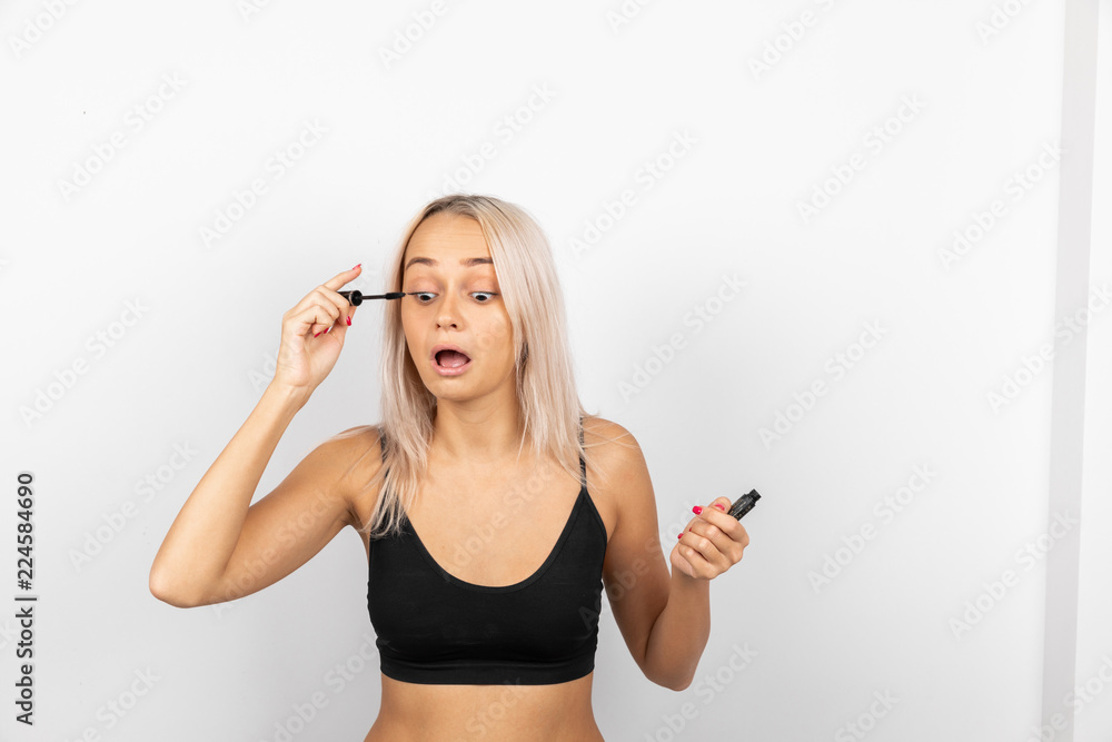 A slender girl with blond hair in a black T-shirt having opened her mouth paints her eyelashes with black mascara on a white background A slender girl with blond hair in a black T-shirt having opened her mouth paints her eyelashes with black mascara on a white background