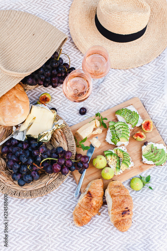 Romantic summer picnic with delicious food, two wineglasses, healthy fruits: avocado, grapes, figs. Straw hat, bag, sandwiches, croissants, cheese are on blanket. Breakfast in italian or french style